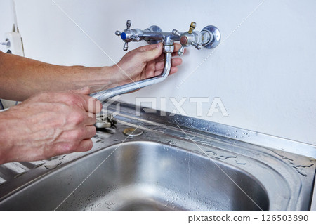 Removing faucet spout from wall-mounted tap by hand in preparation for replacement using visible plumbing tools placed near kitchen sink. 126503890