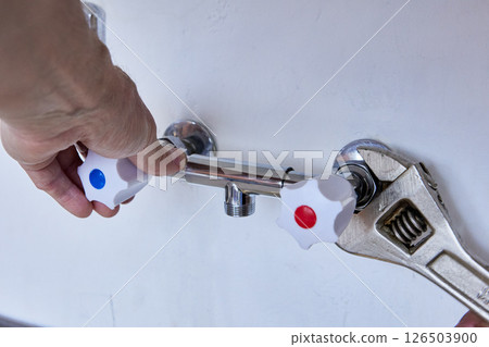 Plumber uses adjustable wrench to secure new two handle wall faucet, one hand holds fixture steady while connections are tightened to plumbing outlets. 126503900