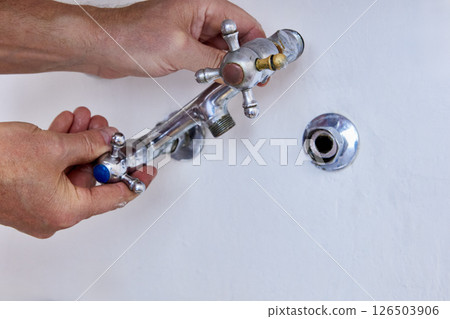 Hands are removing used two handle faucet from wall fitting, worn metal fixture with cross handles and missing spout is separated from exposed connector. 126503906