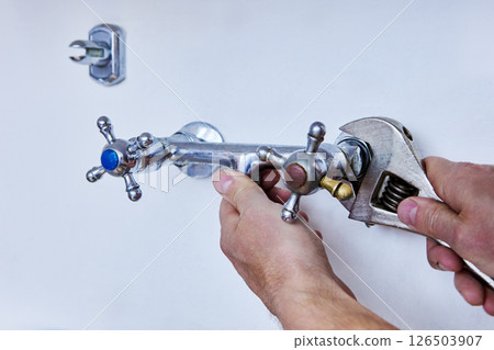 Detaching outdated faucet to install modern fixture, person uses wrench to unscrew metal tap with visible signs of use and colored handle indicators. 126503907