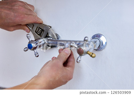 Unscrewing old two handle faucet from wall, hands use wrench to loosen fixture with visible signs of wear, central opening remains without spout. Unscrewing old two handle faucet from wall, hands use wrench to loosen fixture with visible signs of wear, central opening remains without spout. 126503909