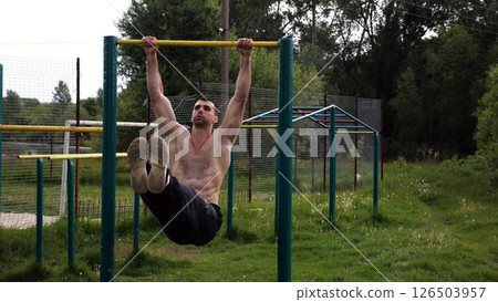 Athletic man trains abdominal muscles raising legs on horizontal bar at sports ground. Young sportsman exercising on pull-up bar outdoor. Sporty athlete working out at nature. Healthy lifestyle 126503957