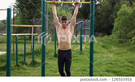 Muscular athlete doing pull ups on horizontal bar at sports ground. Young shirtless sportsman exercising at nature. Strong sporty man training outdoor. Concept of healthy and active lifestyle. Slow mo 126503958