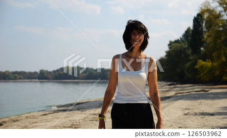 Happy adult woman going on sandy beach near shoreline at sunny day. Beautiful lady walking on seashore with calm waves at background. Concept of summer vacation or holiday. Slow motion 126503962