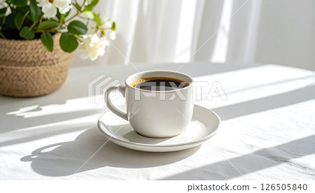 A white coffee cup and saucer on a sunlit table with a plant and curtains in the background. A white coffee cup and saucer on a sunlit table with a plant and curtains in the background. 126505840