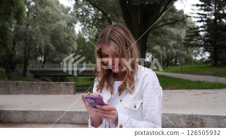 A woman enjoys her time in a park, smiling as she interacts with her smartphone while sitting on a stone surface amid trees and pathways. 126505932
