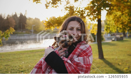 A woman embraces her small dog, both cozy under a blanket, enjoying a warm autumn afternoon by the lake in a picturesque park with vibrant foliage. 126505935