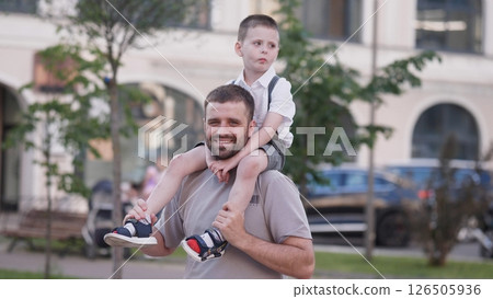 A man joyfully carries his son on his shoulders during a sunny afternoon at the park, surrounded by greenery and urban architecture. A man joyfully carries his son on his shoulders during a sunny afternoon at the park, surrounded by greenery and urban architecture. 126505936