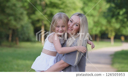 A smiling mother holds her young daughter in a sunny park. They enjoy a playful moment, surrounded by trees and fresh grass on a warm afternoon. 126505953