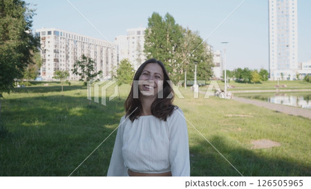 A woman stands in a grassy park area, smiling broadly while enjoying the sunny weather. Nearby, modern buildings and trees enhance the urban landscape. A woman stands in a grassy park area, smiling broadly while enjoying the sunny weather. Nearby, modern buildings and trees enhance the urban landscape. 126505965