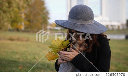 A woman embraces her small dog while holding colorful autumn leaves. Bright sunlight filters through the trees in a vibrant city park during the fall season. A woman embraces her small dog while holding colorful autumn leaves. Bright sunlight filters through the trees in a vibrant city park during the fall season. 126505978