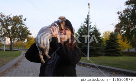A joyful woman holds a small dog in her arms, smiling widely as they enjoy a beautiful autumn day in a serene park filled with colorful foliage. A joyful woman holds a small dog in her arms, smiling widely as they enjoy a beautiful autumn day in a serene park filled with colorful foliage. 126505979
