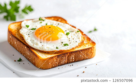 Fried egg on toasted bread with pepper and herbs, served on white paper on a bright surface. Fried egg on toasted bread with pepper and herbs, served on white paper on a bright surface. 126507096