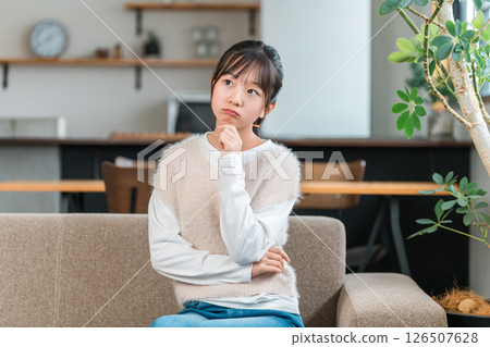 A worried elementary school student or junior high school student girl sits on the sofa in the living room and thinks. A worried elementary school student or junior high school student girl sits on the sofa in the living room and thinks. 126507628