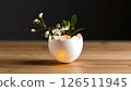 A delicate eggshell vase holds white flowers and leaves, glowing softly on a wooden table against a dark background. 126511945