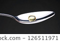A single yellow softgel capsule sits on a silver spoon against a dark background, focusing on health and medicine. 126511971