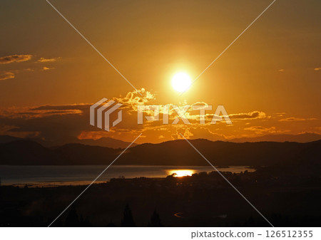 The setting sun shining on the opposite shore of Lake Inawashiro. The orange sky and the sparkling lake surface are impressive and beautiful. The setting sun shining on the opposite shore of Lake Inawashiro. The orange sky and the sparkling lake surface are impressive and beautiful. 126512355