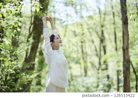 A woman stretches with one arm raised in a forest of fresh greenery. 126513422