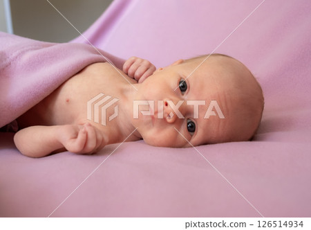 Newborn baby lying on a soft pink blanket while looking curiously at the camera in a cozy indoor setting Newborn baby lying on a soft pink blanket while looking curiously at the camera in a cozy indoor setting 126514934