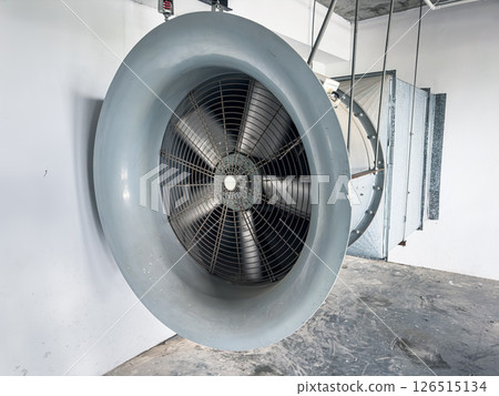 A large, gray, industrial fan is suspended from the ceiling of a concrete building. A large, gray, industrial fan is suspended from the ceiling of a concrete building. 126515134
