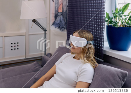 Eye massage device. Young girl wearing glasses for massage relaxing on sofa at home. Woman takes care of her health and skin care 126515886