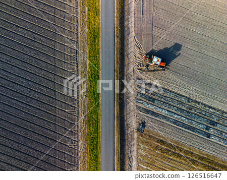 Aerial view of a tractor plowing a field next to a road, with striking patterns in the soil and clear geometric lines dividing the farmland. 126516647