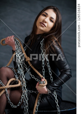 A stunning young woman with long dark hair poses with thick chains in a dramatic studio setting, wearing a tight black leather outfit and conveying a powerful, slightly rebellious mood A stunning young woman with long dark hair poses with thick chains in a dramatic studio setting, wearing a tight black leather outfit and conveying a powerful, slightly rebellious mood 126516834
