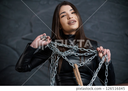 A young Caucasian woman with long brown hair in a black leather outfit playfully holds a thick silver chain and rope against a textured dark gray wall A young Caucasian woman with long brown hair in a black leather outfit playfully holds a thick silver chain and rope against a textured dark gray wall 126516845