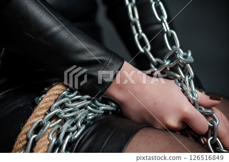 A close-up shot depicts a person's hand bound with ropes and heavy metal chains, evoking a sense of captivity and restriction; the leather clothing suggests a dark aesthetic 126516849