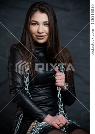 A striking young Caucasian woman with long dark hair poses confidently in a black leather outfit, holding a silver chain, embodying strength and allure 126516850