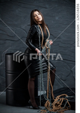 Dramatic portrait of a confident woman in a black leather dress posing with heavy chains against a dark stone wall and industrial barrels, embodying power and resilience Dramatic portrait of a confident woman in a black leather dress posing with heavy chains against a dark stone wall and industrial barrels, embodying power and resilience 126516856