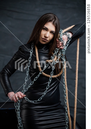 A striking young woman with a determined expression, bound by silver chains and natural rope in a dark, atmospheric setting, wearing a sleek black leather dress A striking young woman with a determined expression, bound by silver chains and natural rope in a dark, atmospheric setting, wearing a sleek black leather dress 126516858