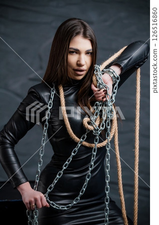 Captivating young woman in black leather dress with thick rope and silver chains, bound hands, intense gaze, dark and moody studio portrait 126516860