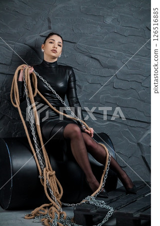 Dark and captivating portrait of a young woman in a black latex dress seated on a modern chair with ropes and chains, embodying strength and mystery Dark and captivating portrait of a young woman in a black latex dress seated on a modern chair with ropes and chains, embodying strength and mystery 126516885