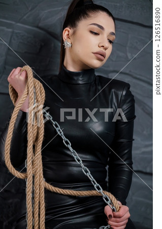 Captivating woman in a sleek black leather outfit poses with a thick rope and silver chain against a dark textured wall, exuding confidence and mystery Captivating woman in a sleek black leather outfit poses with a thick rope and silver chain against a dark textured wall, exuding confidence and mystery 126516890
