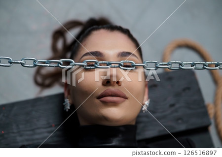 A striking close-up portrait of a young woman with a silver chain draped across her eyes, evoking themes of restraint and confinement, set against a dark, textured backdrop 126516897