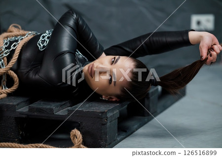 A striking woman with dark hair lies bound on a wooden pallet, secured with ropes and chains in a dimly lit, industrial setting, exuding a captivating yet vulnerable aura A striking woman with dark hair lies bound on a wooden pallet, secured with ropes and chains in a dimly lit, industrial setting, exuding a captivating yet vulnerable aura 126516899
