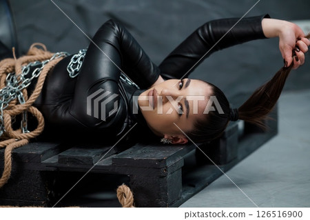 A young woman, seemingly of East Asian descent, lies bound on a wooden pallet with ropes and chains, exhibiting a pensive expression, leather attire, and restrained pose 126516900