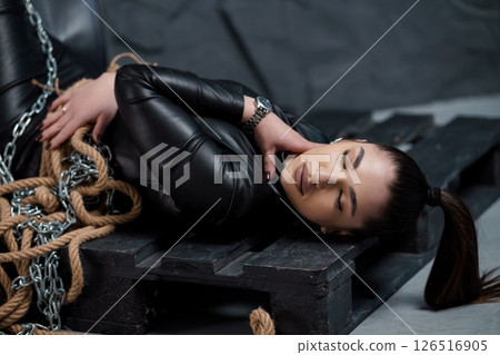 A young Caucasian woman in a black leather suit is bound with chains on a dark wooden pallet, evoking themes of vulnerability and captivity in a dimly lit setting 126516905