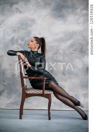 Elegant woman in a black leather dress sitting confidently on a wooden chair against a gray textured backdrop, exuding sophistication and poise in a professional setting 126516926