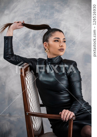 A captivating portrait of a young woman with a sleek ponytail, striking a pose with confidence in a black leather dress, seated on a wooden chair 126516930
