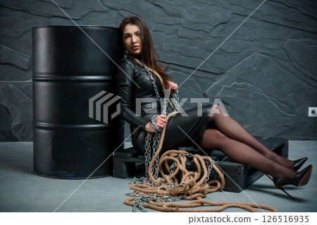 Dramatic portrait of a young woman in a black leather dress chained to a barrel in a dark, textured room, creating a sense of captivity and tension 126516935