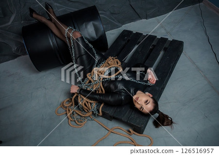 A dramatic scene depicting a young woman bound with rope and chains on a wooden pallet near industrial barrels in a dimly lit warehouse setting, evoking themes of captivity and vulnerability A dramatic scene depicting a young woman bound with rope and chains on a wooden pallet near industrial barrels in a dimly lit warehouse setting, evoking themes of captivity and vulnerability 126516957