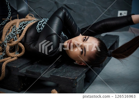 A captivating young woman with brunette hair in a black leather bodysuit is posed lying on a wooden pallet with chains and ropes suggesting a bondage theme in a dark warehouse A captivating young woman with brunette hair in a black leather bodysuit is posed lying on a wooden pallet with chains and ropes suggesting a bondage theme in a dark warehouse 126516963