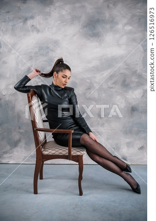 A stylish woman with olive skin and dark hair is seated elegantly in a vintage chair, wearing a black leather dress and sheer black stockings, posing against a textured gray wall 126516973