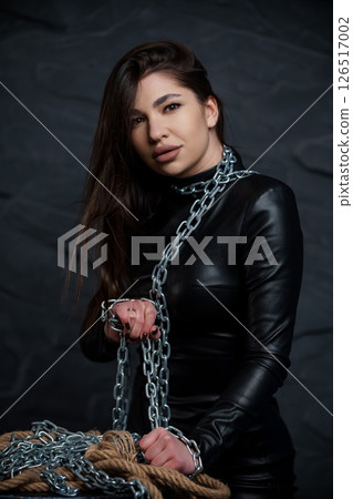 Intricate portrait of a young woman with dark hair wearing a black leather outfit and a silver chain, posing with a confident expression against a dark background Intricate portrait of a young woman with dark hair wearing a black leather outfit and a silver chain, posing with a confident expression against a dark background 126517002