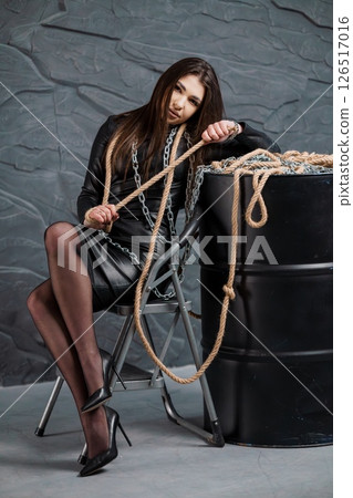 A striking young woman with long brown hair sits on a metal chair, confidently holding a thick rope and a heavy chain, posed against a rough stone wall and a dark barrel A striking young woman with long brown hair sits on a metal chair, confidently holding a thick rope and a heavy chain, posed against a rough stone wall and a dark barrel 126517016