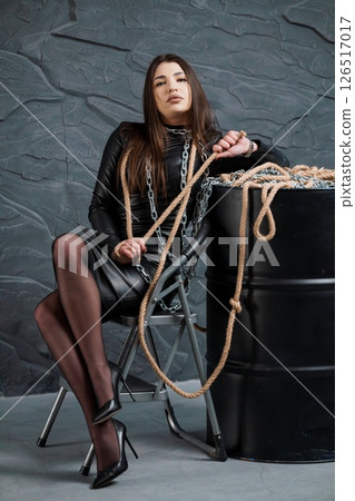 A striking young woman with long dark hair sits confidently on a metal chair, holding a thick rope and chain against a dark stone wall background 126517017