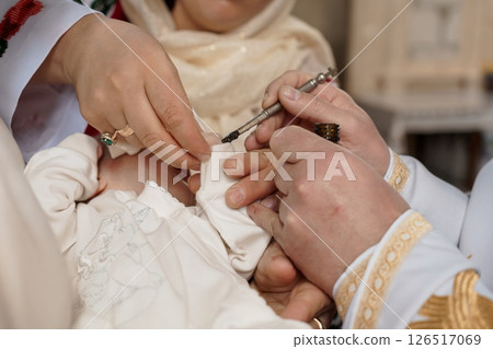 A close-up view of a priest performing the Sacrament of Baptism on a baby, anointing them with chrism oil using a small brush during a religious ceremony 126517069