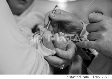 A close-up black and white photograph depicts a solemn baptism ceremony a priest anoints the foot of a newborn baby with holy oil using a small silver vessel signifying spiritual initiation 126517072